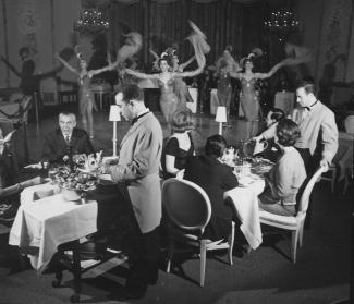 Intérieur de cabaret avec spectacle sur la scène, couples attablés et serveurs.