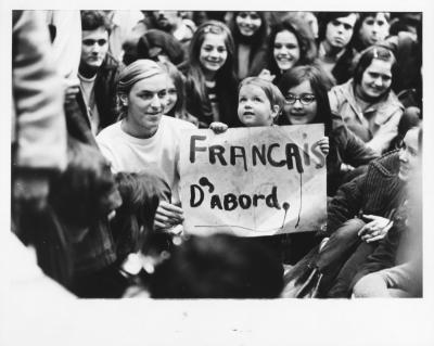 Photographie noir et blanc montrant une foule. Au centre de l’image, deux femmes soutiennent un enfant qui tient une pancarte sur laquelle il est écrit « Français d’abord ».
