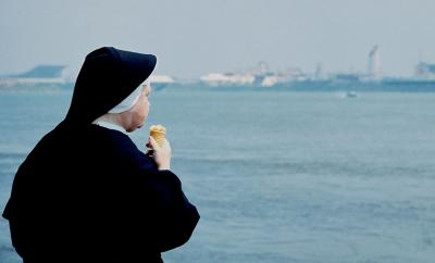 Photographie en couleur montrant une religieuse, de dos, le voile dans le vent mangeant un cornet de crème glacée à la vanille. 