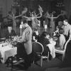 Intérieur de cabaret avec spectacle sur la scène, couples attablés et serveurs.